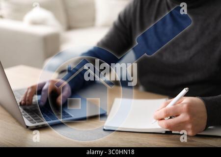 Immobilienmakler mit Laptop und Schreiben in Notizbuch am Tisch, Nahaufnahme. Lupe mit Haussymbol auf dem virtuellen Bildschirm Stockfoto