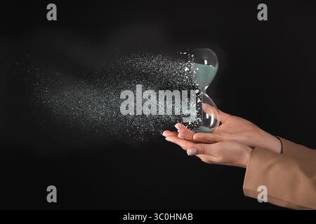 Die Zeit wird knapp. Sanduhr löst sich in den Händen der Frau vor schwarzem Hintergrund auf, Nahaufnahme Stockfoto