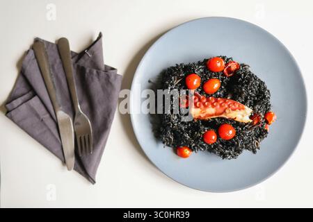 Schwarzes Gourmet-Risotto mit gegrilltem Oktopus und Kirschtomaten auf einem grauen Teller, Feinschmeckergericht aus nächster Nähe. Stockfoto