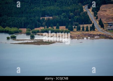 Blick aus der Vogelperspektive auf einen ruhigen See, umgeben von üppigen grünen Wäldern und einer gewundenen Straße. Das Wasser reflektiert die Bäume und den Himmel, in dem eine kleine Insel sichtbar ist Stockfoto