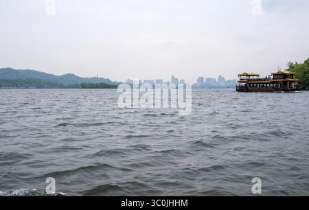 Traditionelles Boot auf dem Westsee, Hintergrund der Skyline von Hangzhou City, China. Nebliger Hintergrund, regnerischer Tag Stockfoto