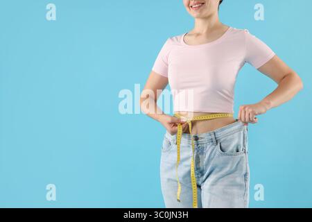 Frau mit Maßband, die alte Jeans nach Gewichtsverlust auf hellblauem Hintergrund trägt, Nahaufnahme. Leerzeichen für Text Stockfoto