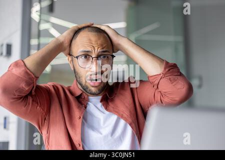 Ein besorgter, junger Mann mit Brille reagiert auf das, was er auf seinem Laptop sieht, und hält seinen Kopf in einem besorgten Zustand. Stockfoto