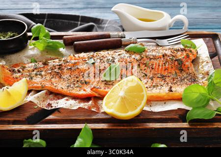 Gegrillte ganze Lachsfilets mit Haut auf einem rustikalen Holzschneidebrett mit frischem Basilikum, Zitronenscheiben, Besteck und Gewürzen auf Holztisch, Horizo Stockfoto