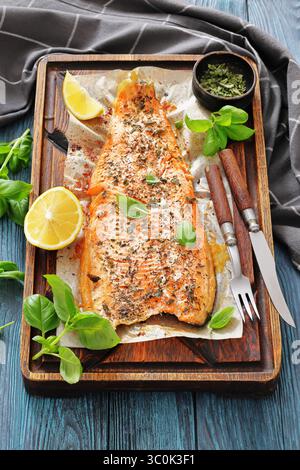 Gegrillte ganze Lachsfilets mit Haut auf einem rustikalen Holzschneidebrett mit frischem Basilikum, Zitronenscheiben, Besteck und Gewürzen auf Holztisch, vertica Stockfoto