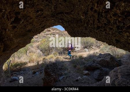 Weibliche Wanderer, die am Eingang einer vulkanischen Höhle in einer trockenen Schluchtlandschaft vorbeispazieren Stockfoto