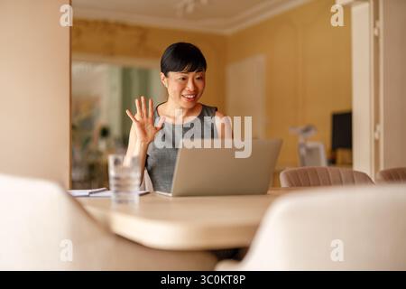 Glückliche Geschäftsfrau winkt Hand, während sie online über Videoanrufe auf einem Laptop im Firmenbüro diskutiert Stockfoto