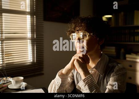 Kaukasische Frau mittleren Alters, die am Schreibtisch sitzt und das Kinn auf den Händen liegend und nachdenklich in die Ferne blickt, mit einer Brille, die durch das Fenster im Hintergrund strahlt Stockfoto