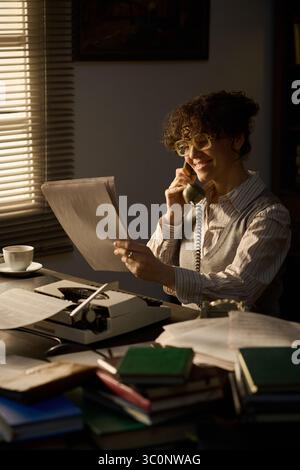Kaukasische junge Erwachsene Frau, die am Schreibtisch sitzt und mit einem Festnetztelefon spricht, während sie Dokumente in der Hand hält, umgeben von Büchern und Papieren, lächelt und im Büro mit der Schreibmaschine arbeitet Stockfoto