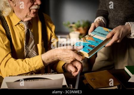 Ein älterer kaukasischer Mann, der an der Rezeption sitzt und ein Buch von einer jungen, erwachsenen kaukasischen Frau erhält. Beide konzentrieren sich auf den Austausch eines bunten Reisebuches mit dem Titel My Journey Stockfoto