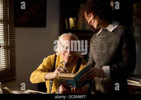 Ein älterer kaukasischer Mann sitzt am Schreibtisch und unterschreibt ein Buch, während eine junge Erwachsene kaukasische Frau neben ihm lächelt. Beide arbeiten freundlich im Büro zusammen Stockfoto