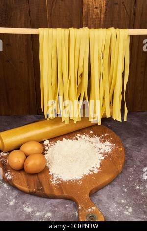 Frische Eifettuccinnudeln, im Vordergrund trocknen, über einem Holztisch mit Mehl und Ei hängen, Grundzutaten für die Zubereitung. Stockfoto