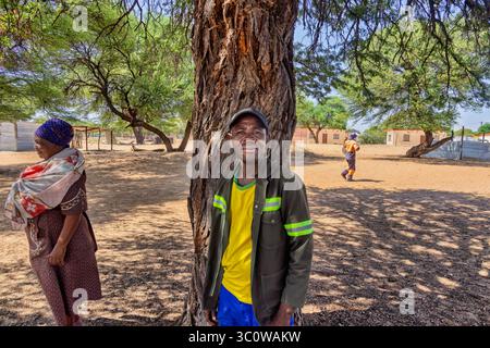 Dorf, Porträt eines lächelnden alten Mannes in afrika, im Schatten eines Baumes, Häuser im Hintergrund, vorbeiziehende Menschen und umstehende Personen Stockfoto