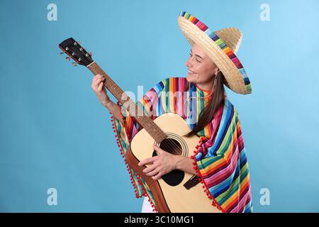 Lächelnde Frau in Sombrero-Hut und farbenfrohem Poncho, der Gitarre auf hellblauem Hintergrund spielt Stockfoto