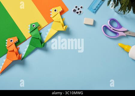 Origami Arbeitstisch mit Papier, Googly Eyes, Schere, Kleberflasche, Lineal, und Radiergummi auf hellblauem Hintergrund. Fertige Dinosaurierspielzeuge aus Papier. Leerzeichen f Stockfoto