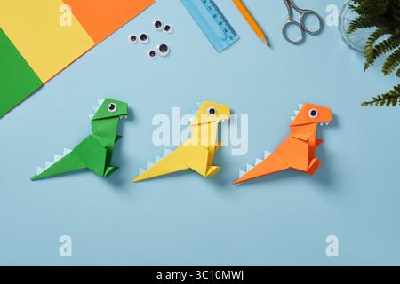 Origami-Arbeitstisch mit Papierbögen, guten Augen, Schere, Kleberflasche, Lineal, und Radiergummi auf hellblauem Hintergrund. Fertiges Dinosaurierspielzeug aus Papier Stockfoto