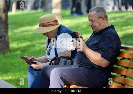 Moskau, Russland - 18. Juli 2025: Altes Ehepaar sitzt auf einer Bank im Sommerpark. Mann und Frau, die Smartphones benutzen, Leben im Ruhestand Stockfoto