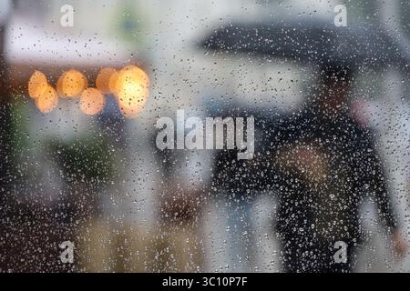 Starker Regen in einer Stadt, unscharfer Blick durch nasses Glas mit Wassertropfen für Leute, die mit Sonnenschirmen laufen Stockfoto
