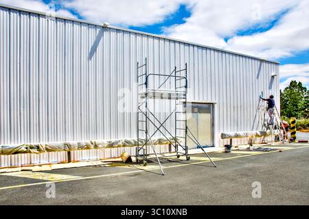 Die Außenwände eines Lagerhauses aus geriffeltem Metall werden von Bauunternehmern neu gestrichen - Chatellerault, Vienne (86), Frankreich. Stockfoto