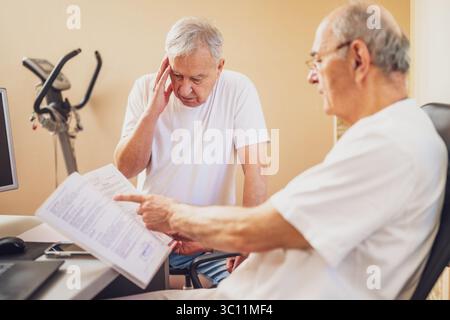 Patient wird beim Kardiologen untersucht. Der Doktor sieht sich seine medizinischen Akten an. Der Patient beschwert sich über Schmerzen. Stockfoto