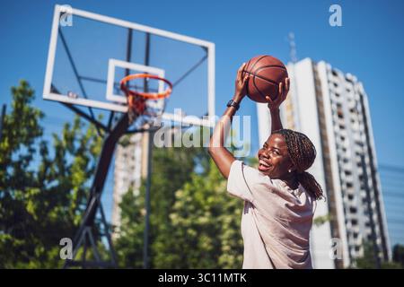 Junge Frau, die morgens alleine Basketball auf dem Sportplatz übt. Sie schaut in die Kamera und lächelt. Stockfoto
