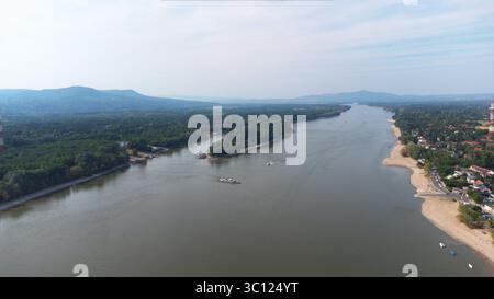 Drohnenvideo, Donau in der Nähe von Budapest, die sich durch eine üppige, bewaldete Region mit Sandstrand und fernen Hügeln schlängelt. Boote auf dem Wasser, kleine Flüsse Stockfoto
