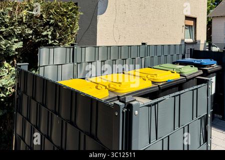 Mehrere Recyclingbehälter in verschiedenen Farben sind ordentlich hinter einem Wohngebäude angeordnet. Die Abfalleimer sind von einem hohen Zaun umschlossen, wodurch ein aufgeräumtes W Stockfoto