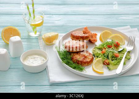 Geräucherte Lachsburger, Pasteten oder Kuchen, serviert mit Krautsalat und Zitronenscheiben auf einem weißen Teller auf Holztisch mit Tzatziki-Sauce und Sprudelwasser, Stockfoto