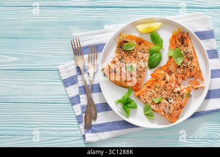 Gegrillte ganze Lachsfilets mit Haut auf einem Teller mit frischem Basilikum, Zitronenscheiben, Gabeln auf Holztisch, horizontale Ansicht von oben, flache Lage, frei Stockfoto