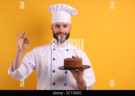 Glücklicher Konditor in Uniform, der Schokoladenkuchen hält und eine gute Geste auf orangem Hintergrund zeigt Stockfoto