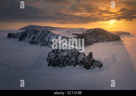 Aus der Vogelperspektive erheben sich die schneebedeckten Klippen des Kap Khoby aus der gefrorenen Weite des Baikalsees unter einem feurigen Sonnenuntergangshimmel, Russland. Stockfoto