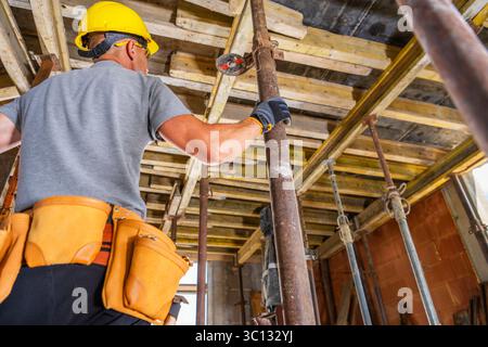 Ein Bauarbeiter mit gelbem Schutzhelm und grauem Hemd stellt ein Gerüst in einem Gebäude auf. Werkzeuge und Materialien sind um ihn herum sichtbar, während er arbeitet. Stockfoto