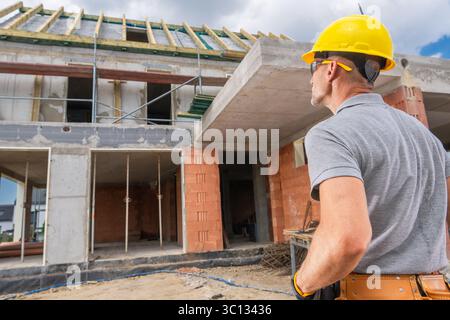 Bauarbeiter mit gelbem Schutzhelm beobachtet, wie Arbeiter bei Tageslicht mit Ziegeln und Beton ein modernes Gebäude bauen. Stockfoto