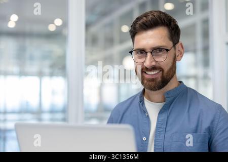 Ein lächelnder, bärtiger Mann mit Brille schaut direkt in die Kamera. Er trägt ein blaues Hemd und ist im Büro. Stockfoto