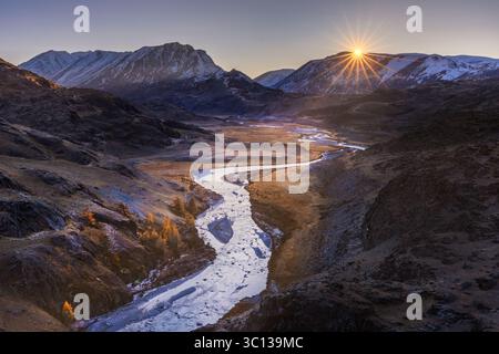 Blick auf einen sich windenden Fluss, der sich durch ein zerklüftetes Tal schlängelt, goldenes Licht, das die schneebedeckten Gipfel küsst, während die Sonne über dem Horizont bricht, Altai Republik, Russland. Stockfoto