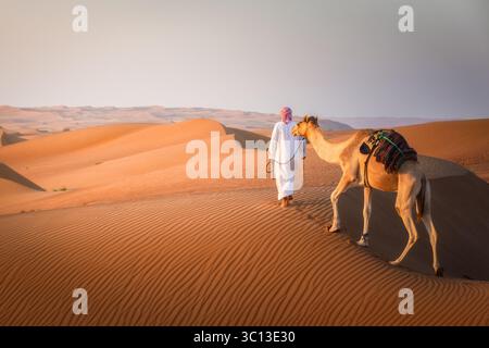 Wahiba Sands, Oman – 12. Mai 2022: Blick auf einen Mann in traditionellen weißen Gewändern, der ein Kamel über die hügeligen, sonnengeküssten Dünen führt und lange Schatten auf den welligen Sand wirft. Stockfoto