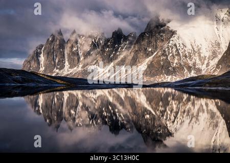 Blick auf zerklüftete, schneebedeckte Gipfel, die sich im stillen Wasser spiegeln, eine Symphonie aus Grau und weiß unter einem brütenden Himmel, Tungeneset, Norwegen. Stockfoto