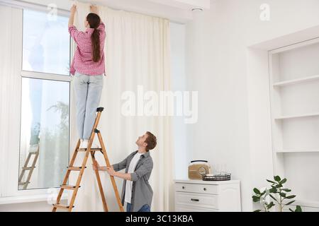 Ein Paar, das Vorhänge mit einer Leiter im Zimmer aufstellt Stockfoto