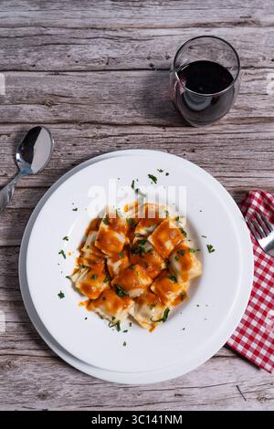 Köstliche Ravioli-Pasta mit lebendiger Tomatensauce, bestreut mit frischer Petersilie, serviert auf einem weißen Teller, begleitet von einem Glas Rotwein auf Rost Stockfoto