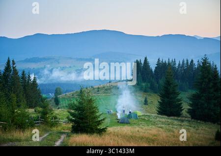Campingplatz aus der Vogelperspektive in den Bergen, mit mehreren bunten Zelten auf einer grasbewachsenen Rodung. Camper entspannen unter klarem blauen Himmel, umgeben von hohen immergrünen und Panoramablick auf entfernte Gipfel. Stockfoto