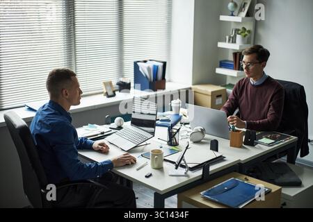 Zwei kaukasische junge Erwachsene Männer, die an Schreibtischen in modernen Büros arbeiten, beide mit Laptops und gegenüberliegend, diskutieren über das Projekt mit Dokumenten und Bürobedarf auf dem Tisch Stockfoto