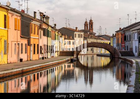 Blick auf lebendige, farbenfrohe Häuser entlang eines ruhigen Kanals unter einer bogenförmigen Brücke, Reflektionen im Wasser, Comacchio, Emilia-Romagna, Italien. Stockfoto