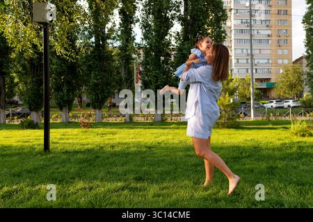 Barfuß-Mutter in gestreiften Shorts hebt und dreht ihre kichernde Tochter in einem blauen Kleid über hellem Gras und teilt fröhliche Sommermomente neben Bäumen A Stockfoto