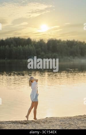 Eine barfuß Frau läuft am Sandstrand des Waldsees entlang. Sie setzte ihr Gesicht auf die warmen Strahlen der Sonnenuntergangssonne Stockfoto