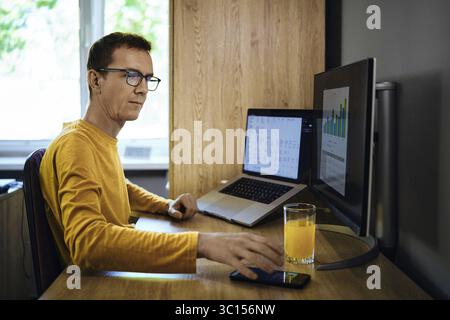Ein Mann in einem gelben Hemd sitzt an einem Schreibtisch in einem Heimbüro. Er wurde durch einen Anruf vom Studium der Statistik abgelenkt Stockfoto