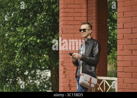 Ein Mann in Lederjacke und Sonnenbrille steht an roten Ziegelsäulen. Er hält ein Smartphone und sucht nach einem nahenden Taxi Stockfoto