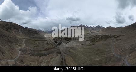 Aus der Vogelperspektive schlängelt sich eine gewundene Straße durch das zerklüftete, trockene Gelände in Richtung schneebedeckter Berge unter einem bewölkten Himmel, Leh, Ladakh, Indien. Stockfoto