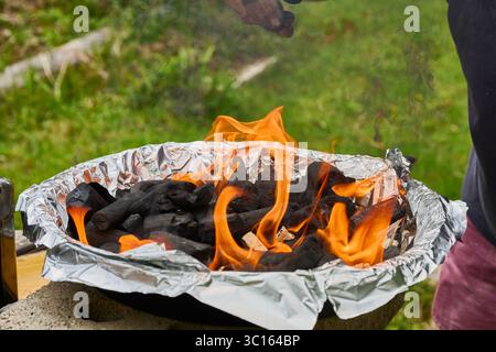 Holzkohle brennt in Flammen in einem tragbaren Grill, der mit Aluminiumfolie bedeckt ist und für das Grillen im Freien bereit ist Stockfoto