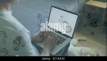 Arbeitender Mann in weißem T-Shirt und Jeans, der mit Laptop im Schlafzimmer studiert, mit Hund ausgeruht Stockfoto