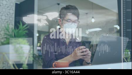 Smartphone-Mann, der Maske und Brille trägt, analysiert Karten am Schreibtisch, mit Laptop Stockfoto